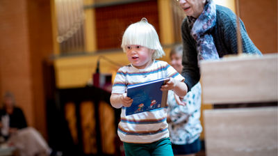 Gutt løper med 4årsbok i hendene. Han er inne i et kirkerom og det sitter andre rundt. Gutten smiler.