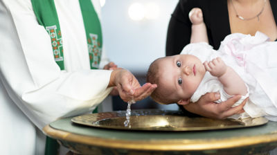 Det er dåp i kirken. Et barn blir holdt over en døpefont, og skal til å få vann over hodet.
