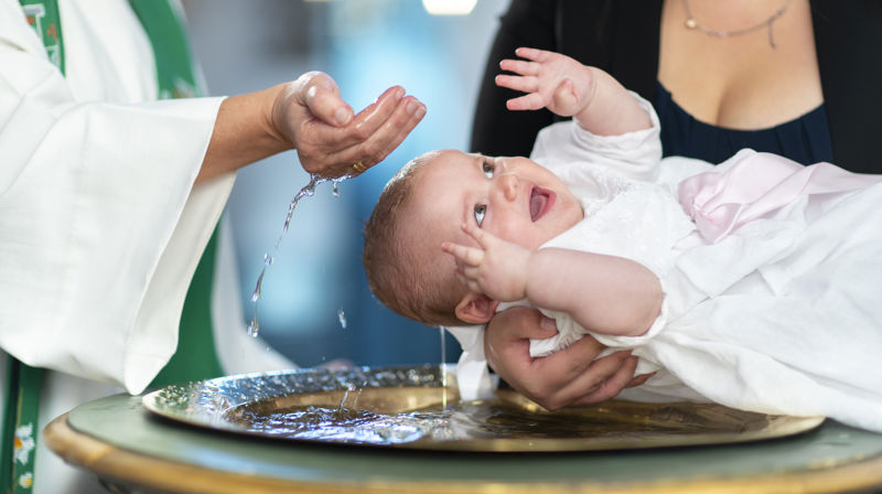 Det er dåp i kirken. Et barn blir holdt over en døpefont, og skal til å få vann over hodet.