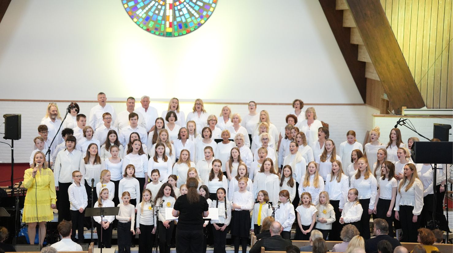 Stort barne- og ungdomskor kledd i hvitt med solist og dirigent. Foto.