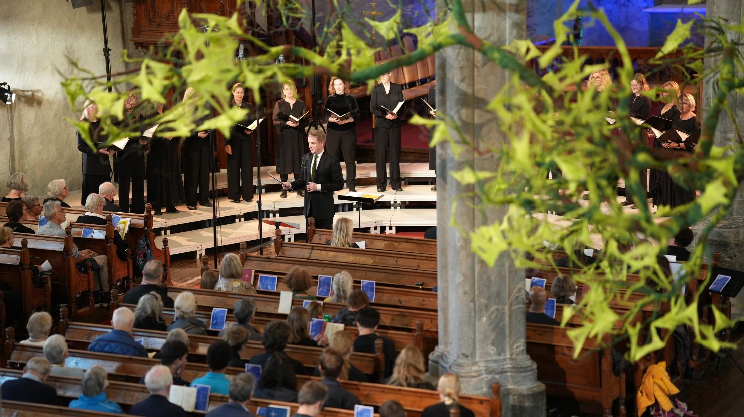 Korkonsert i kirke, folk i benker og damekor på scenen. Dirigenten snakker. Foto.