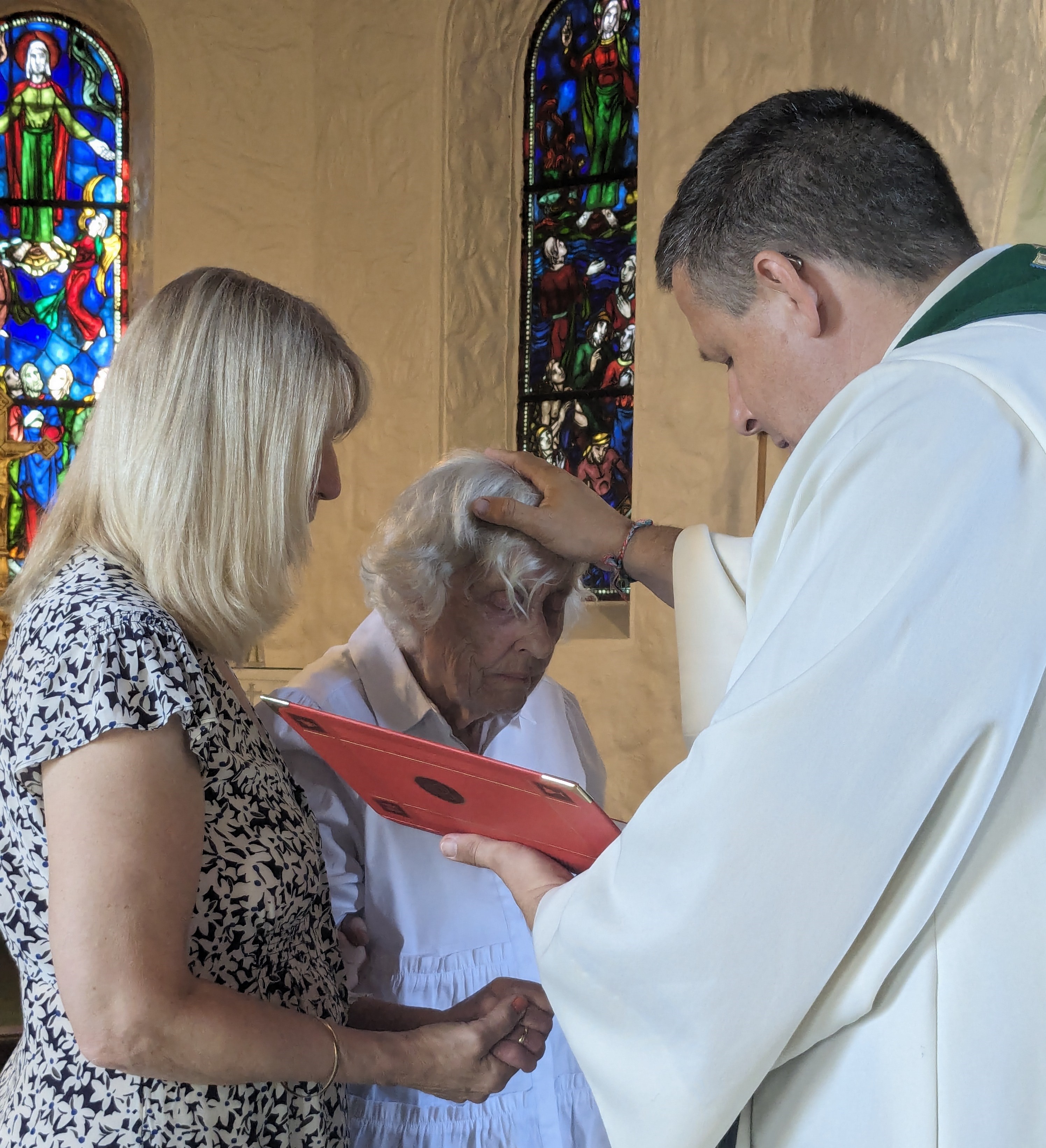 Bildet viser tre personer, en eldre dame som står over døpsfonten, en prest som holder håndet over hodet hennes og velsigner henne, samt en dame som holder den eldre damen i hånden.