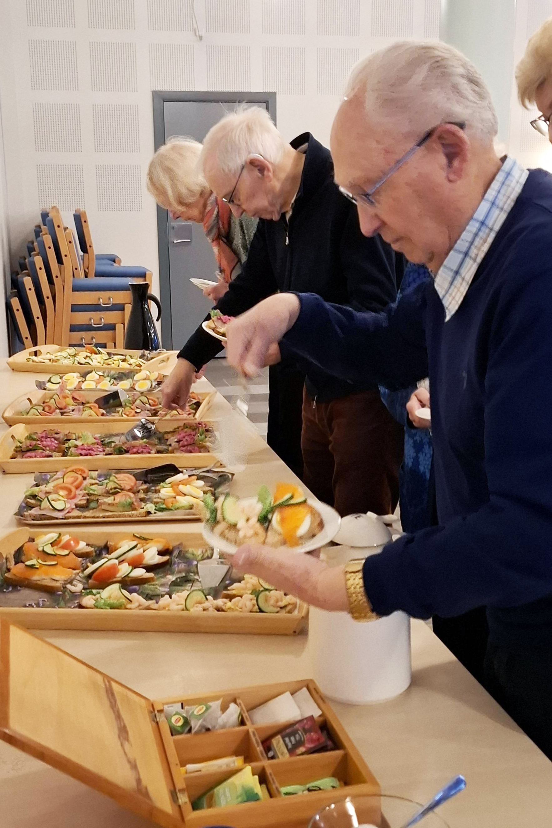 Tre eldre står og forsyner seg av mat som er lagt ut på et langbord.