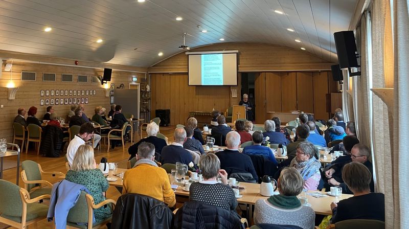 Årsmøte i kyrkjelydssalen, mange mennesker sitter på stoler rund bordene og følger med på en presentasjon på lerretet fremst i salen.