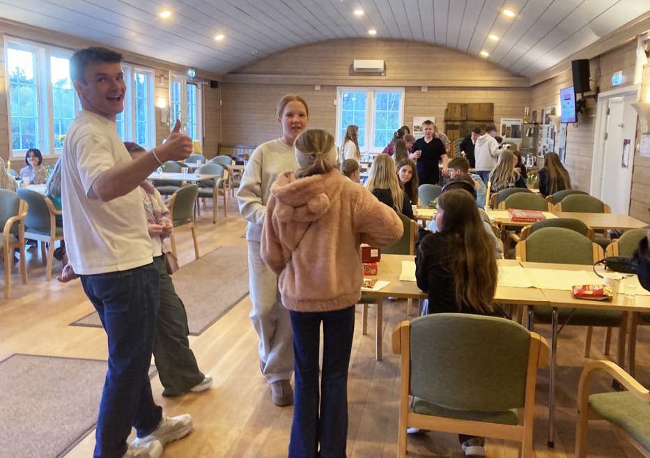 Ungdommer og tweensere sitter og står spredt inne i kyrkjelydssalen, til høyre står en mann og gliser og holder tommel opp