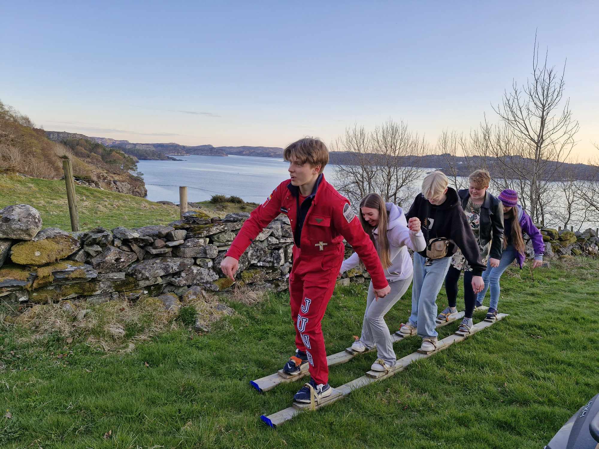 Fem ungdommar står på to plankar, festa på beina som ski, og balanserer bortover graset