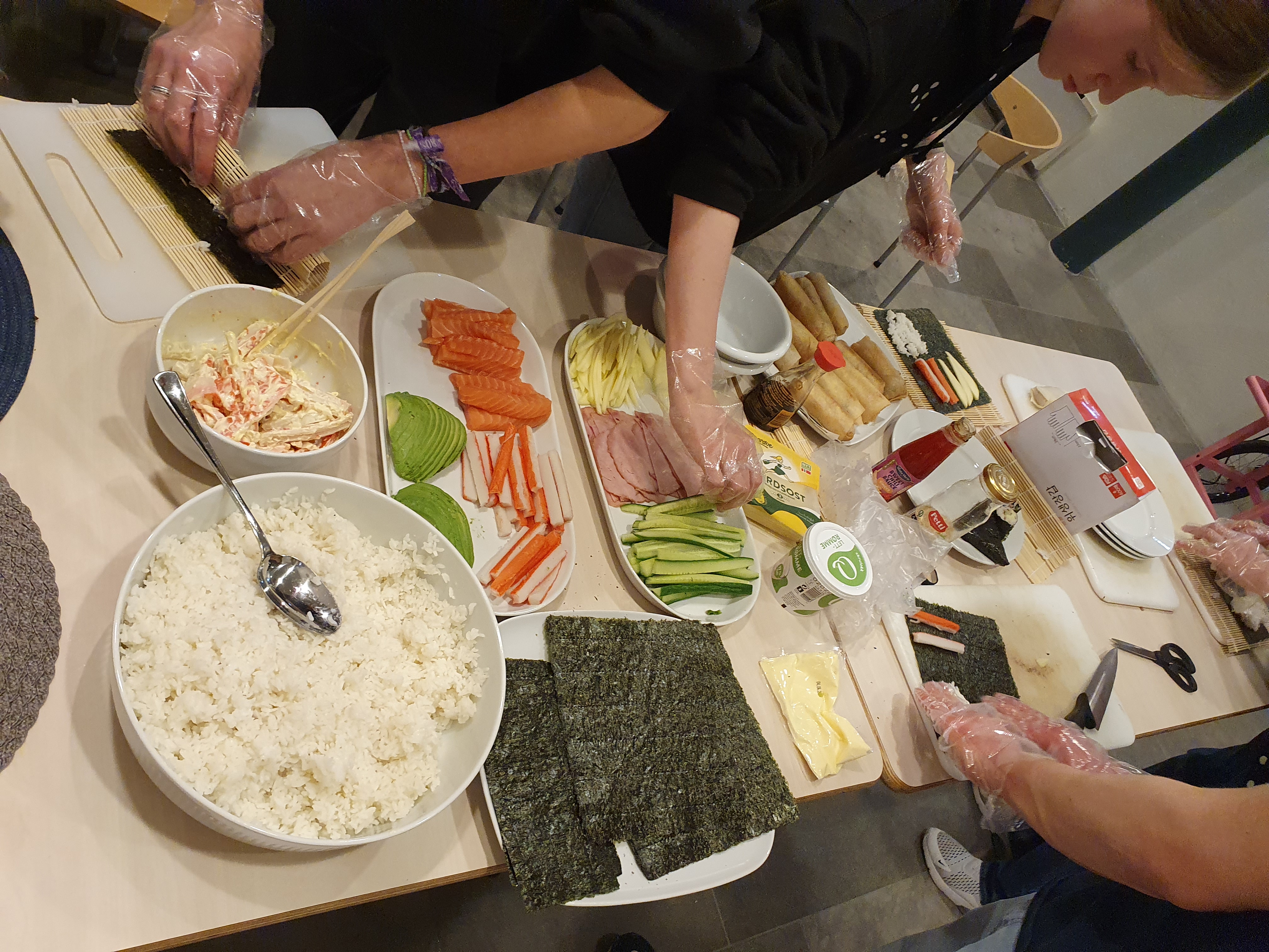 Ungdommar står ved eit bord, dei har plasthanskar på og lager og ruller sushi. Ulike ingredienser ligg på bordet.