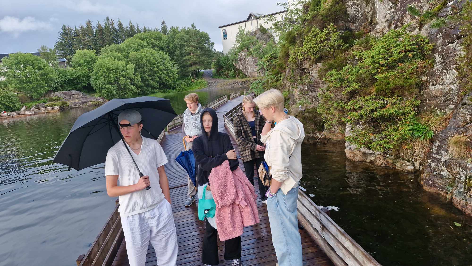 Ungdommar står på brygga nedanfor Foldnes kyrkje, ein har paraply, og ein annan har eit handkle