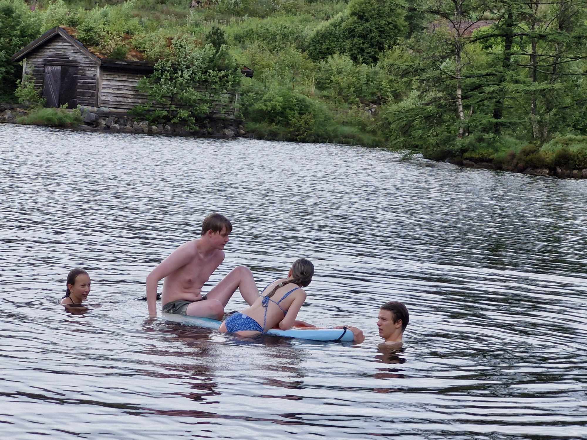 Fire ungdommar badar i vatnet, to av dei lenar seg/sit på eit brett.