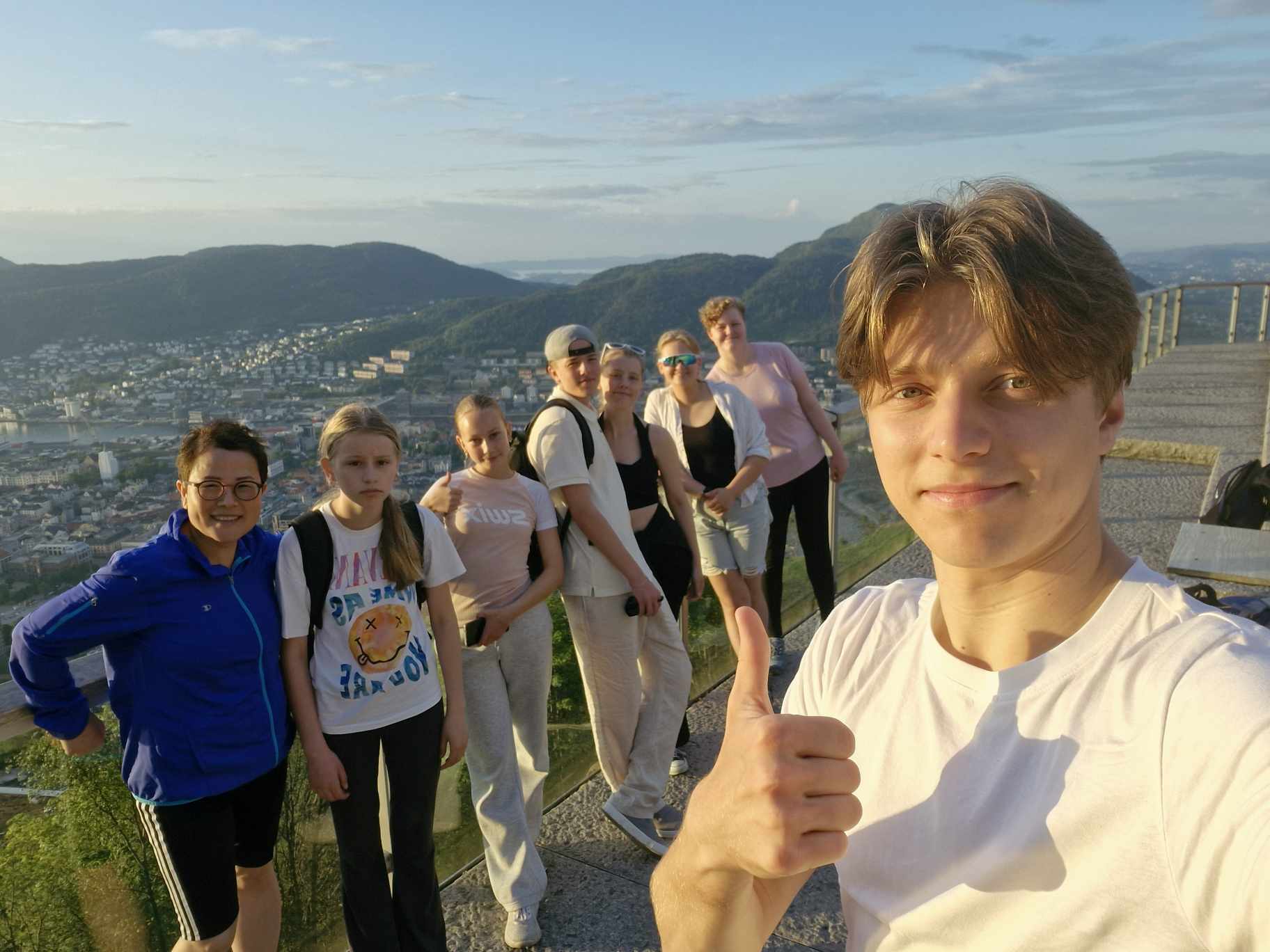 Ein ungdom tek selfie med tommelen opp, bak står fleire ungdommar og smiler til kameraet. Dei er oppe på Føyen og vi ser Bergen i bakgrunnen.