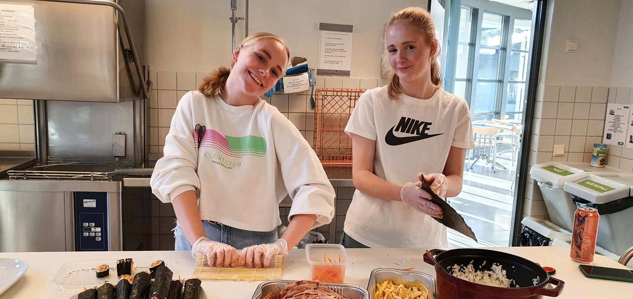 To ungdomsjenter står på kjøkkenet og lager mat, de smiler til kameraet