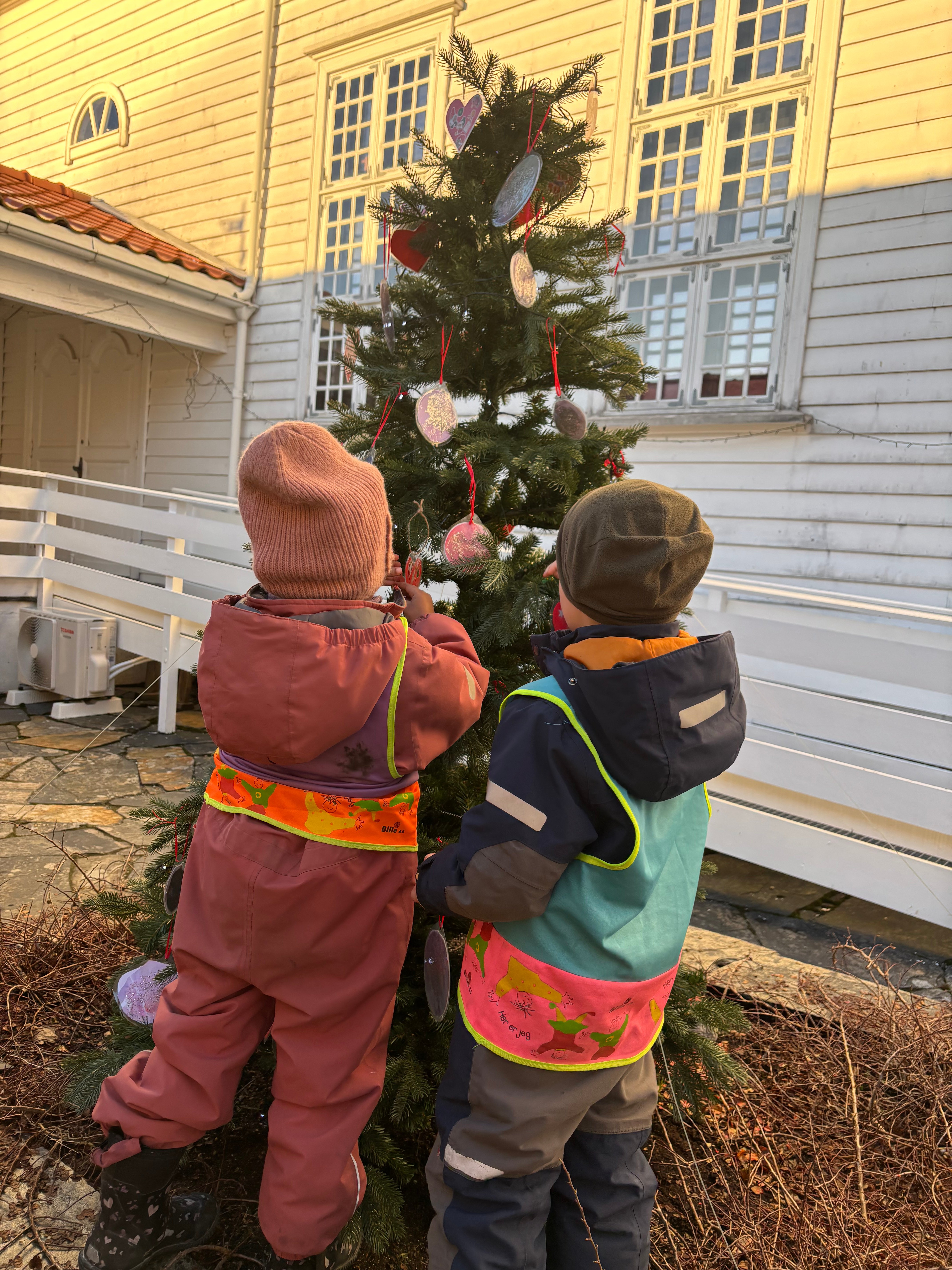 To barnehagebarn står i borggården og henger pynt opp et lite juletre