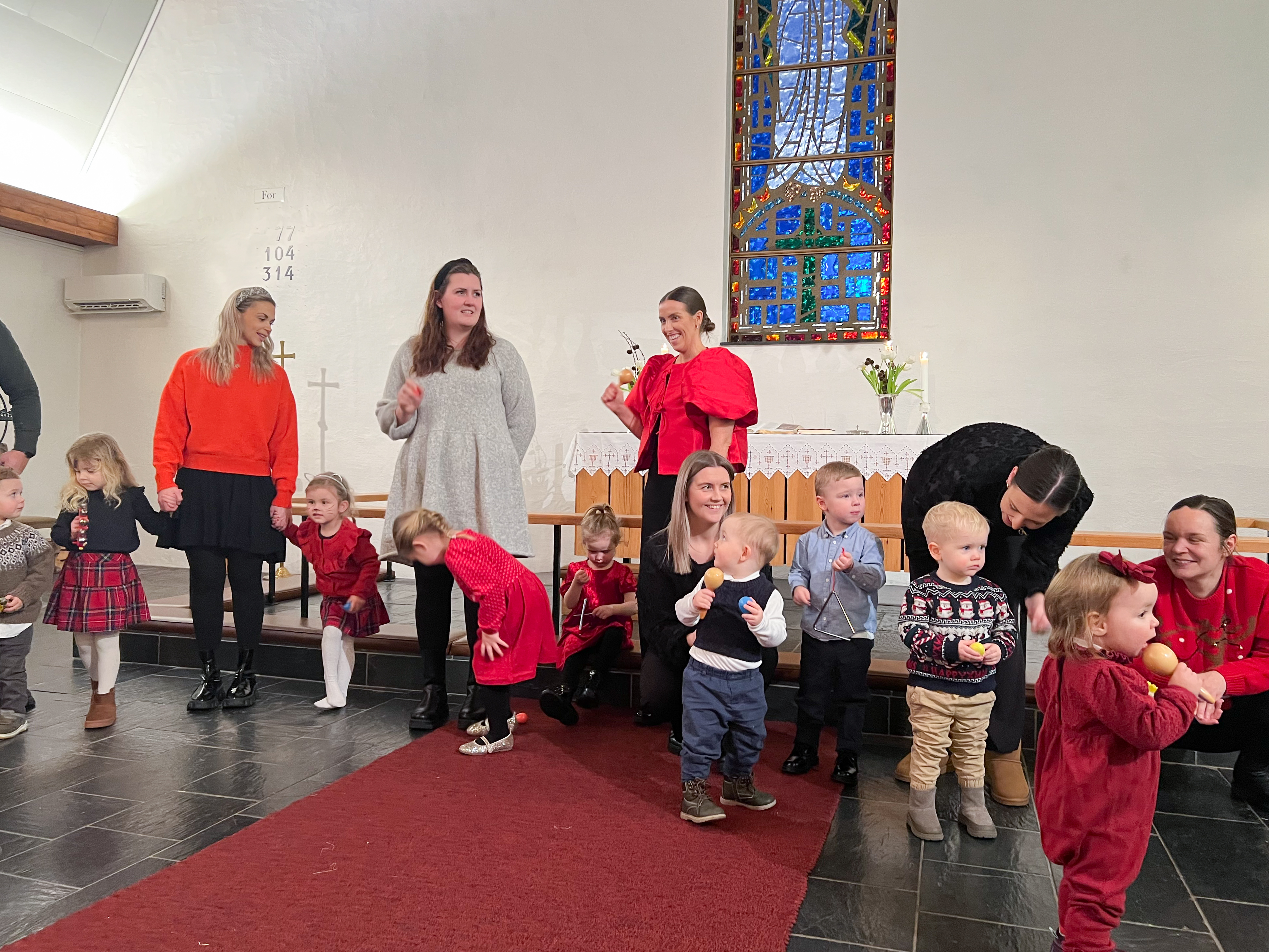 Små barn og mødre står fremme i hjelme kirke og synger med rasleinstrumenter i hendene