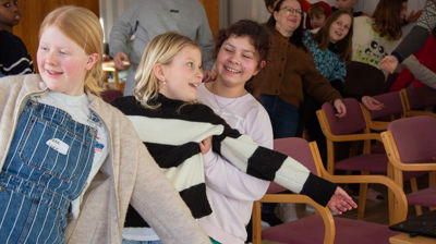 Tre jenter står og lener seg bak mot hverandre og smiler. Man kan se andre barn, voksne og ungdommer i stolrader bak de i samme bevegelse.