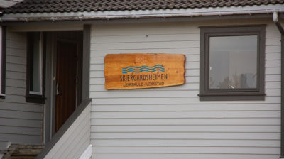 Bilde av en hvit yttervegg av et hus, på veggen henger det et skilt av tre med Skjergardsheimen sin logo skjert inn.