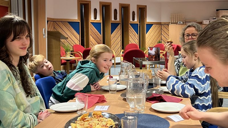 Barn og voksne sitter rundt et bord og spiser pizza, en jente lener seg over bordet og smiler