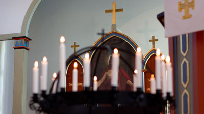 Utsnitt av korset over altertavlen i kirken, med tente lys i lysgloben uklart i forgrunnen