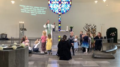Presten står framme i kyrkja saman med mange 4-åringar og nokre foreldre, ho lagar bevegelser til ein song